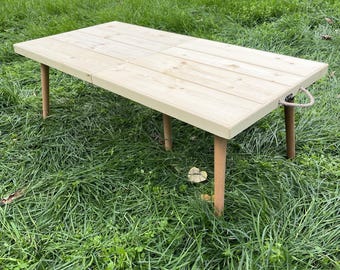May include: A light-colored wooden folding table with four legs. The tabletop is made of wooden planks. The legs are a light brown color. A rope handle is attached to the side of the table. The table is set on a grassy surface.