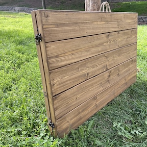 Puede incluir: Una mesa plegable de madera con una mancha marr&oacute;n c&aacute;lida. La mesa est&aacute; construida con tablones de madera horizontales y presenta bisagras de metal negro. Un asa de cuerda natural es visible en la parte superior. La mesa est&aacute; sobre una superficie cubierta de hierba.