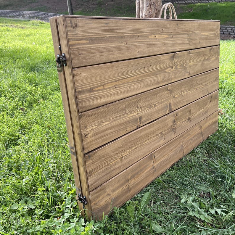 Puede incluir: Una mesa plegable de madera con una mancha marr&oacute;n c&aacute;lida. La mesa est&aacute; construida con tablones de madera horizontales y presenta bisagras de metal negro. Un asa de cuerda natural es visible en la parte superior. La mesa est&aacute; sobre una superficie cubierta de hierba.