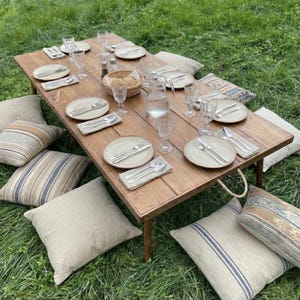 May include: A low, rectangular wooden picnic table set for a meal, surrounded by pillows on the grass. The table is set with plates, silverware, glasses, and a basket. The table is approximately 48 inches long.