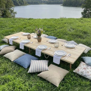 Puede incluir: Una mesa de picnic r&uacute;stica de madera preparada para una comida, rodeada de cojines sobre una zona de c&eacute;sped. La mesa est&aacute; puesta con platos blancos, cubiertos y servilletas. Un jarr&oacute;n con flores est&aacute; en el centro, con un lago al fondo.