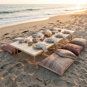 Peut inclure: Une table de pique-nique sur une plage de sable au coucher du soleil. Une table en bois clair est dressée avec des assiettes, des verres et de la nourriture. Des coussins décoratifs sont disposés autour de la table. L'océan est en arrière-plan.