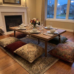 Peut inclure: Une table à manger basse en bois, dressée pour un repas, entourée de coussins de sol à motifs. La table est garnie d'assiettes blanches, de bols, de couverts et de verres. Un arrangement floral et des bougies complètent l'ambiance.
