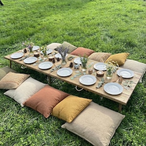 Peut inclure: Une table basse en bois dressée pour un pique-nique sur l'herbe, avec des assiettes blanches, des fleurs et des décorations. La table est entourée de coussins colorés, créant un coin salon confortable pour un rassemblement en plein air.