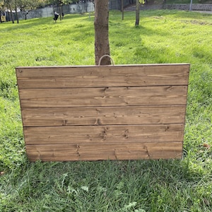 Puede incluir: Una jardinera rectangular de madera con acabado de madera natural. La caja est&aacute; construida con tablones horizontales y tiene un asa de cuerda. La jardinera est&aacute; sobre hierba verde.