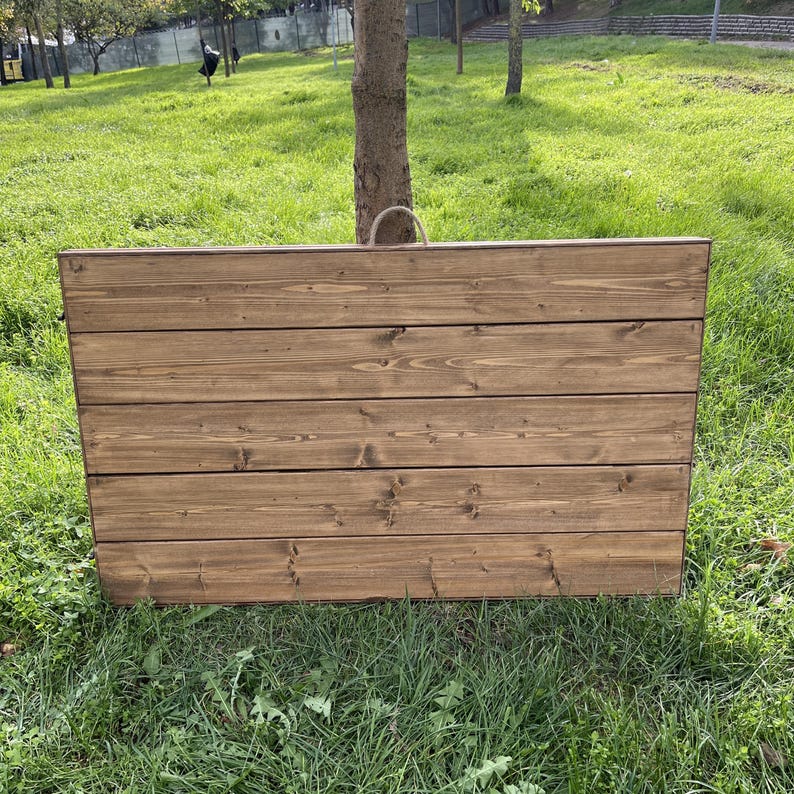 Puede incluir: Una jardinera rectangular de madera con acabado de madera natural. La caja est&aacute; construida con tablones horizontales y tiene un asa de cuerda. La jardinera est&aacute; sobre hierba verde.