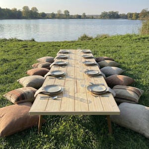 Puede incluir: Una mesa larga de madera clara preparada para una comida al aire libre, rodeada de grandes cojines. Cada cubierto incluye un plato, cubiertos y un vaso. La mesa est&aacute; colocada sobre una zona de c&eacute;sped cerca de un lago.