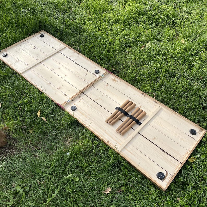 Puede incluir: Un tablero de juego de madera de color claro con forma rectangular. El tablero tiene una serie de clavijas de madera sujetas por una banda el&aacute;stica negra. El tablero est&aacute; sobre una superficie cubierta de hierba.