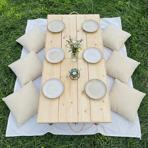 May include: A wooden picnic table with six plates, a small vase of flowers, and a candle holder. Beige pillows surround the table, set on a white blanket over green grass. The table is approximately 30 inches long.