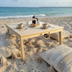 May include: A light-coloured wooden picnic table on a sandy beach, set with coffee, pastries, and teacups. Decorative pillows surround the table. A French press coffee maker and a white teapot are also on the table.