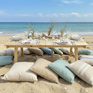 May include: A beach picnic setting with a light wood table set with plates, cutlery, and glasses. Decorative cushions in various colours surround the table. The ocean and blue sky are in the background.