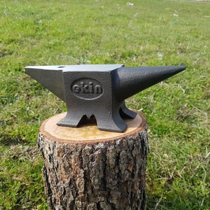 May include: A black anvil with the word "ekin" embossed on the side, resting on a tree stump. The anvil has a flat top, a pointed horn, and a sturdy base. The background is green grass.