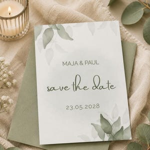 May include: A rectangular save the date card featuring sage green watercolour leaf illustrations, with the text MAJA & PAUL, save the date, and the date 23.05.2028 printed in dark grey, resting on a solid sage green envelope.