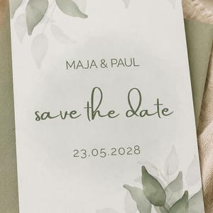 May include: A rectangular save the date card featuring sage green watercolour leaf illustrations in the corners. The text reads MAJA & PAUL, save the date, and 23.05.2028 in a combination of sans-serif and cursive fonts on a white background.