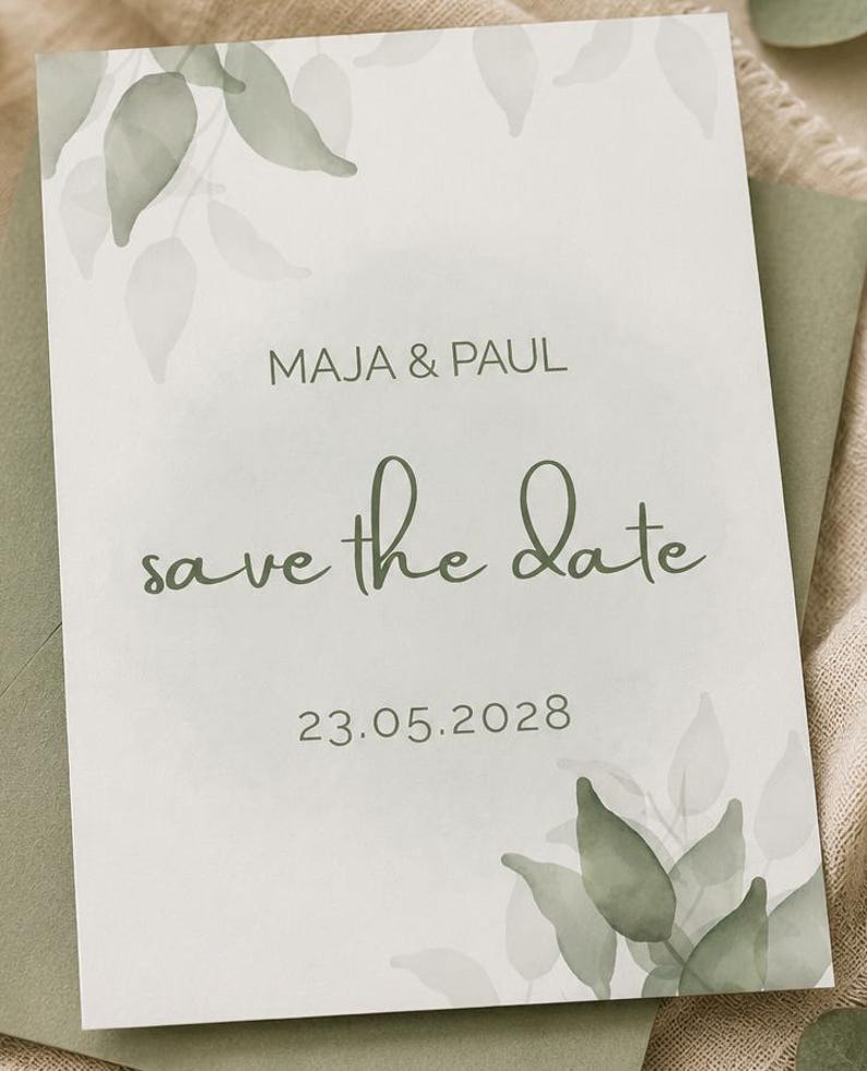 May include: A rectangular save the date card featuring sage green watercolour leaf illustrations in the corners. The text reads MAJA & PAUL, save the date, and 23.05.2028 in a combination of sans-serif and cursive fonts on a white background.