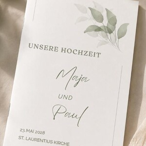 Może przedstawiać: Biała broszura ślubna z akwarelową ilustracją zielonych liści, napisem "UNSERE HOCHZEIT", imionami "Maja" i "Paul" ze słowem "UND" pomiędzy nimi oraz datą "23. MAI 2028" w "ST. LAURENTIUS KIRCHE".