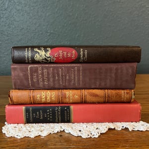 May include: A stack of four vintage books with various colors and textures. The titles include "Dr. Jekyll and Mr. Hyde," "The Pickwick Papers," "The Tragic Comedians," and "The Adventures of Tom Sawyer." The books rest on a white lace doily.