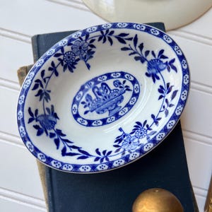 May include: An oval ceramic dish featuring a white base with intricate blue floral patterns and a central basket motif. The rim is decorated with a blue cross-hatch border, and the overall design displays a classic blue and white color scheme.