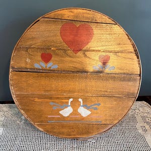 May include: Round wooden decorative piece with a painted design. The piece features a large red heart, two smaller hearts, and two white geese with blue ribbons. The wood has a warm, natural tone.