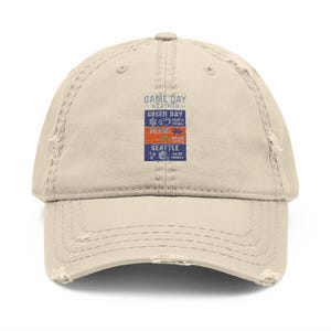 Peut inclure: Casquette de baseball beige délavé avec le texte "GAME DAY WEATHER" au-dessus de trois noms de villes : Green Bay, Miami et Seattle. Le nom de chaque ville est imprimé au-dessus d'un graphique et d'une date. La casquette a une visière incurvée et un aspect usé.