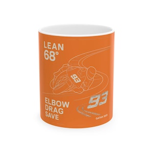 May include: Orange ceramic mug with a white outline of a motorcycle rider leaning into a turn. The text "LEAN 68°" is at the top, and "ELBOW DRAG SAVE" is at the bottom. The number "93" is on the side of the motorcycle and in the bottom right corner. The text "QATAR 2023" is at the bottom.