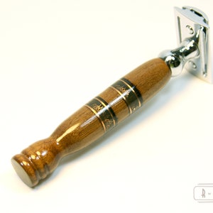 Puede incluir: Una navaja de afeitar de seguridad de madera con una cabeza cromada y un mango de madera marr&oacute;n con incrustaciones doradas y negras.