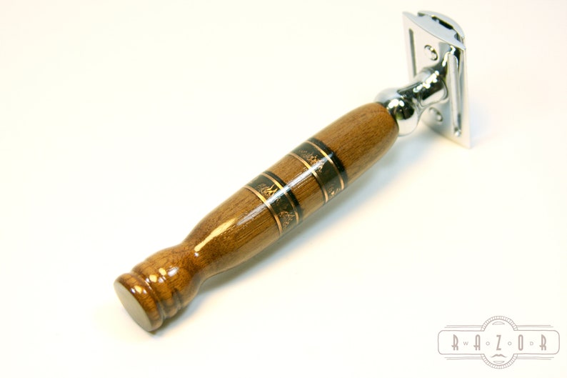 Puede incluir: Una navaja de afeitar de seguridad de madera con una cabeza cromada y un mango de madera marr&oacute;n con incrustaciones doradas y negras.