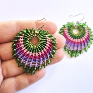 Può includere: Orecchini in macramè fatti a mano a forma di foglia, con un cerchio centrale e un motivo a raggi. Gli orecchini presentano fili verdi, rosa e viola, accentuati da perline verdi. Gli orecchini sono tenuti in una mano.