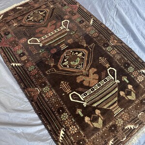 May include: A rectangular, dark brown rug with intricate patterns. The rug features a central design of stylized vases and floral motifs in shades of brown, green, and white. The border has a repeating floral pattern.