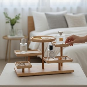 May include: A multi-tiered wooden organizer with round and square trays. The organizer holds perfume bottles, jewelry, and a bracelet. A hand is reaching for a ring on one of the trays. The organizer is on a table with a neutral-colored tablecloth.