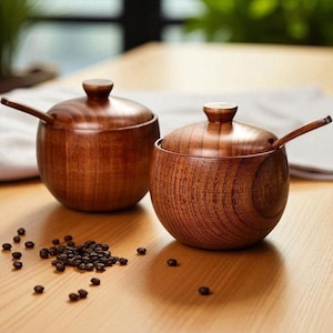 May include: Two wooden spice jars with lids and small spoons. The jars are a warm brown color and sit on a light brown wooden surface. A small pile of dark brown coffee beans is scattered nearby. The jars have a smooth, polished finish.