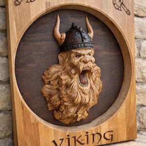 Può includere: Arte da parete in legno raffigurante una testa di vichingo scolpita con un elmo cornuto e la bocca aperta. L'opera è incorniciata con un bordo marrone chiaro, con la parola "Viking" incisa in basso. Due intagli di asce in alto.