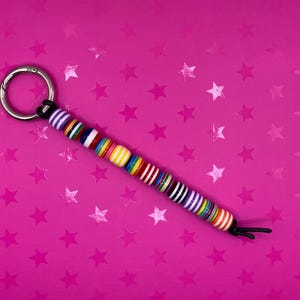 May include: A beaded keyring with a silver metal ring and a black cord. The beads are colourful, with stripes of white, purple, red, yellow, and green. The background is a bright pink with star patterns.