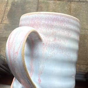 May include: Handmade ceramic mug with a light blue and pink glaze. The mug has a textured, ridged body and a rounded handle. The base is a natural, unglazed colour. This unique mug is perfect for coffee, tea, or any beverage.