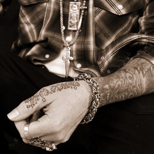 May include: A close-up of a person's arm with a detailed tattoo and a silver chain bracelet. The person is wearing a plaid shirt and has a silver necklace with a cross pendant.