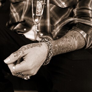 May include: A close-up of a person's arm with a silver chain bracelet and a tattoo on their forearm. The tattoo is in black ink and features a floral design. The person is wearing a plaid shirt and has a silver necklace with a pendant.