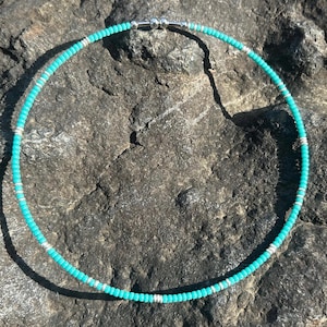 以下が含まれることがあります： シルバーのアクセントが付いた、ターコイズビーズのネックレス。 ネックレスは、小さな丸いターコイズビーズと、シルバーカラーのビーズが交互に連なっています。 ネックレスは、暗く質感のある石の表面に表示されています。