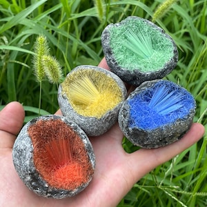 Può includere: Una mano mostra quattro piccoli oggetti grigi simili a rocce, ognuno con un interno di colore diverso. Gli interni sono riempiti con un gruppo di cristalli sottili a forma di ago in verde, giallo, blu e arancione.