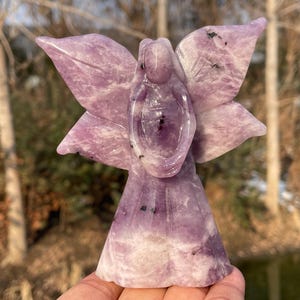 Peut inclure: Une figurine d'ange en améthyste sculptée avec des ailes déployées. L'ange est de couleur violet clair avec des inclusions violet foncé. Les ailes sont détaillées avec un motif en forme de pétale. La figurine est debout.