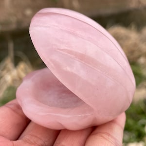 May include: A pink, clam-shaped stone carving, open to reveal a carved interior. The stone has a smooth, polished surface with subtle veining. The clam shell is held open, showcasing the interior. The overall theme is natural and decorative.