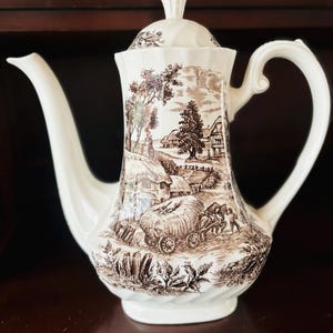 May include: A cream-coloured ceramic teapot featuring a brown transferware print of a rural landscape. The scene depicts a horse-drawn cart loaded with hay, thatched-roof cottages, and trees. The teapot has a curved handle, a long spout, and a lid.