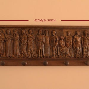 May include: A wooden wall-mounted coat rack with a carved relief of a group of people. The rack is 24.5 inches wide and 7.8 inches tall. It has six hooks for hanging coats, hats, or other items.