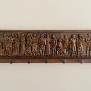 May include: A wooden wall-mounted coat rack with a carved relief of a group of people. The rack has six hooks and a dark brown finish.