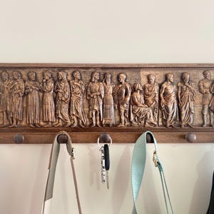 May include: A wooden coat rack with a carved relief of figures in a procession. The rack has multiple hooks for hanging coats, bags, or keys.