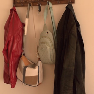 May include: A wooden coat rack with carved figures, featuring three hooks with a red jacket, a brown and white handbag, a light blue crossbody bag, and a dark green jacket hanging from them.