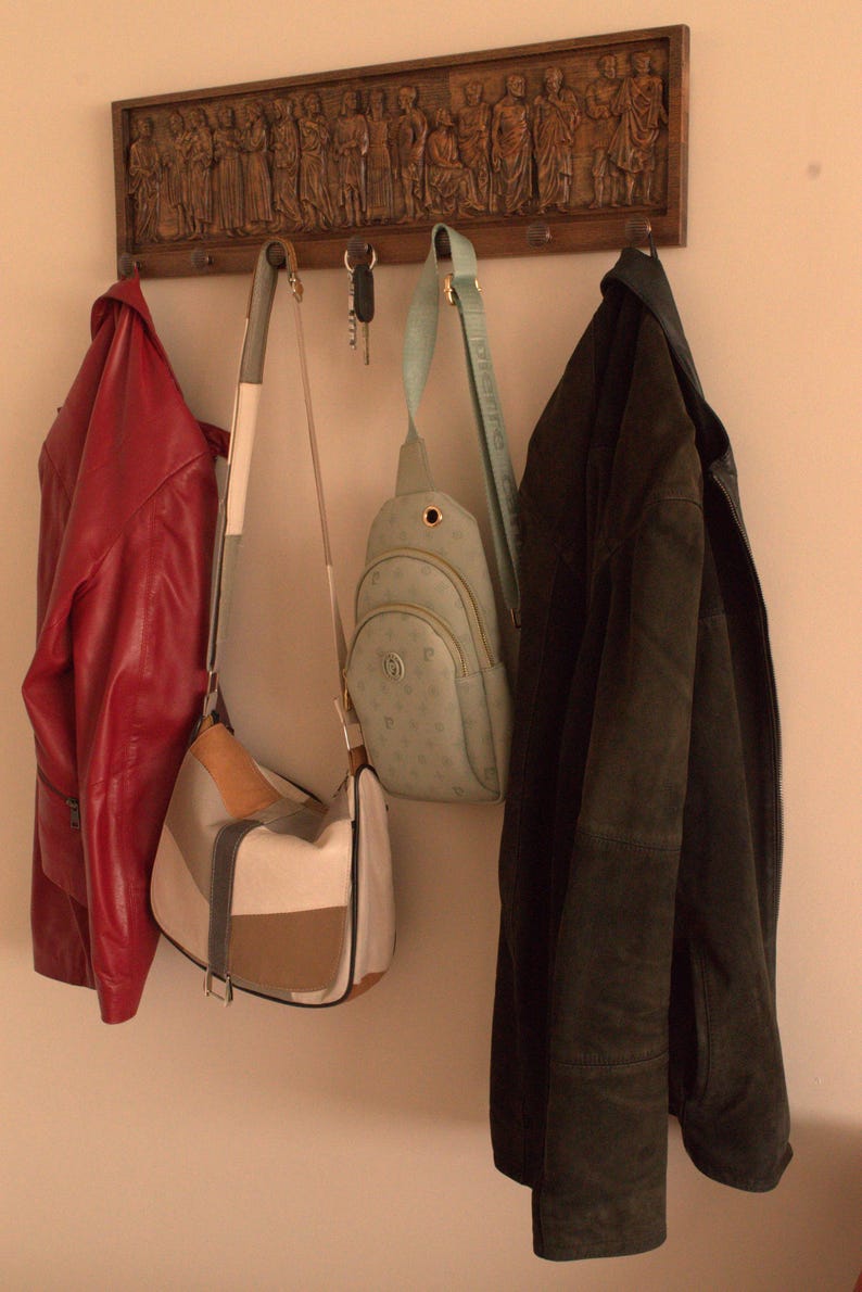 May include: A wooden coat rack with carved figures, featuring three hooks with a red jacket, a brown and white handbag, a light blue crossbody bag, and a dark green jacket hanging from them.