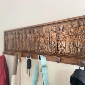 May include: A wooden coat rack with a carved design of figures in togas. The rack has five hooks and is mounted on a white wall. There are three bags hanging on the hooks.