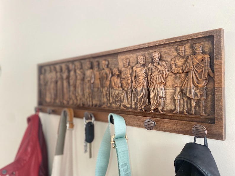 May include: A wooden coat rack with a carved design of figures in togas. The rack has five hooks and is mounted on a white wall. There are three bags hanging on the hooks.