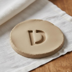 May include: A round, beige ceramic coaster with the letter "D" carved into its surface. The coaster is set on a white linen cloth, with a wooden surface in the background. The coaster is approximately 4 inches in diameter.
