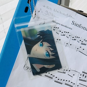May include: A blue binder holds sheet music with a clear plastic sleeve containing an anime character card. The sheet music is labeled "Sinfonia No. 3" and includes handwritten notes. The card features a character with blue eyes.